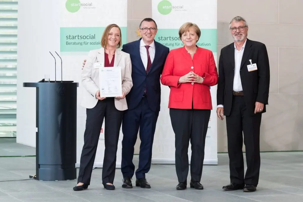 Vier gut gekleidete Personen stehen und schauen in die Blickrichtung der Kamera. Eine Person davon ist die Ex-Bundeskanzlerin Angela Merkel in roten Jacket und eine ist der Initiator des Bundesverbands Burnout und Depression Thomas Grünschläger. Dieses Bild findet man auf der Webseite vom Bundesverband Burnout und Depression.